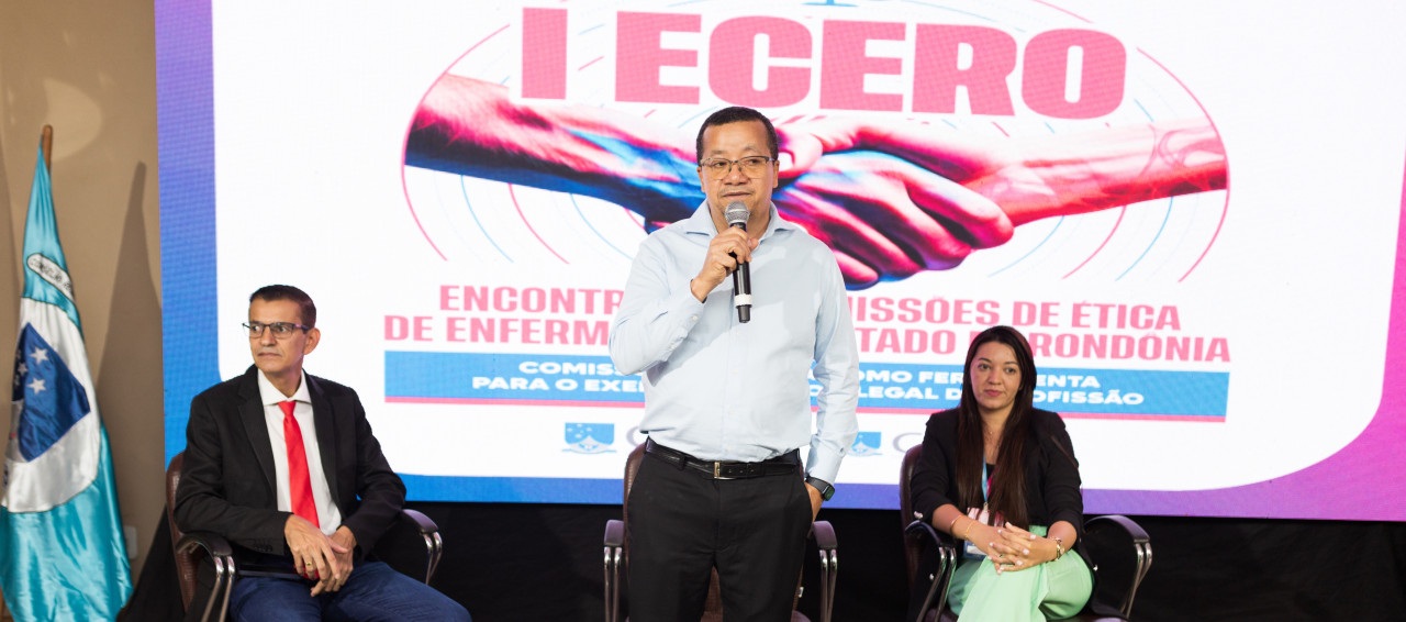 Três pessoas em uma mesa de evento e uma falando ao microfone