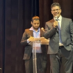 Homem no palco com microfone fazendo alusão a apresentação.