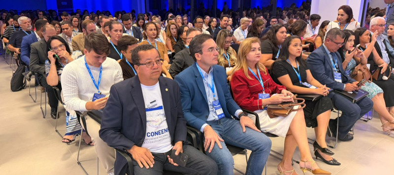 Várias pessoas sentadas em uma sala grande, assisntindo um evento.