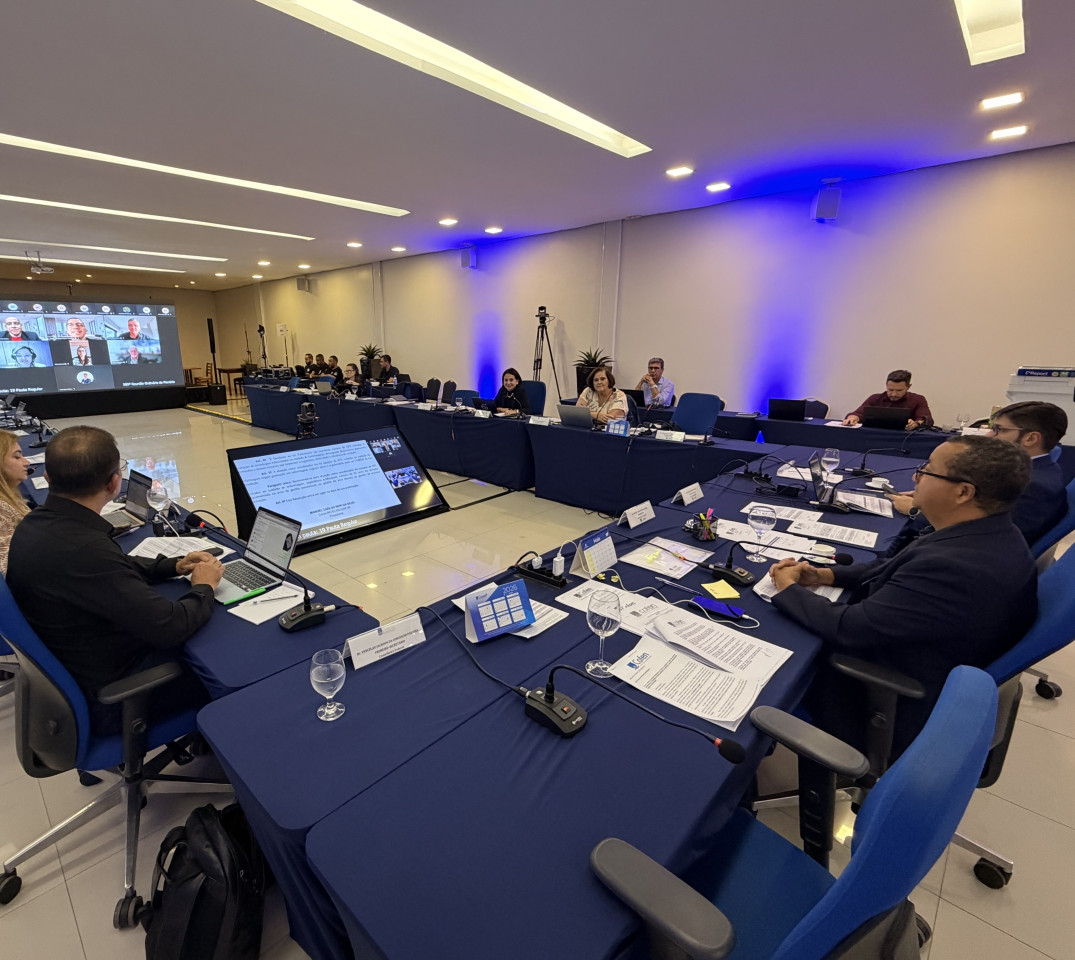 Reunião em sala com homens e mulheres sentados ao redor de uma mesa, enquanto um telão exibe participantes conectados virtualmente.