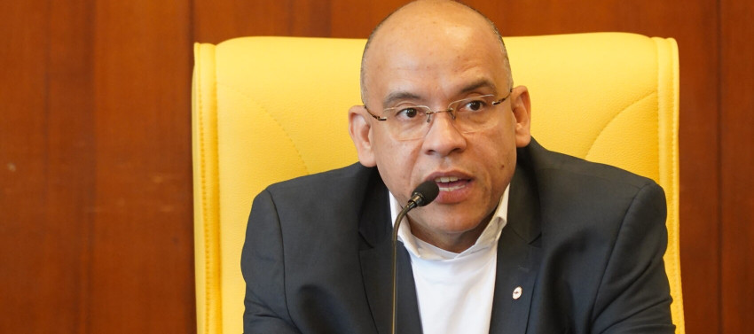 Pessoa sentada na mesa de abertura de uma audiência pública falando no microfone para pessoas