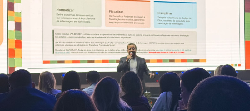 Pessoa na frente do palco de um auditório falando para outras pessoas que estão sentadas